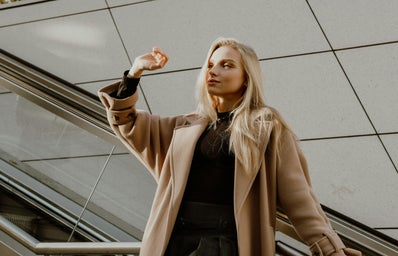 Woman in a camel coloured wool coat, blocking the sun in her face with her hand.