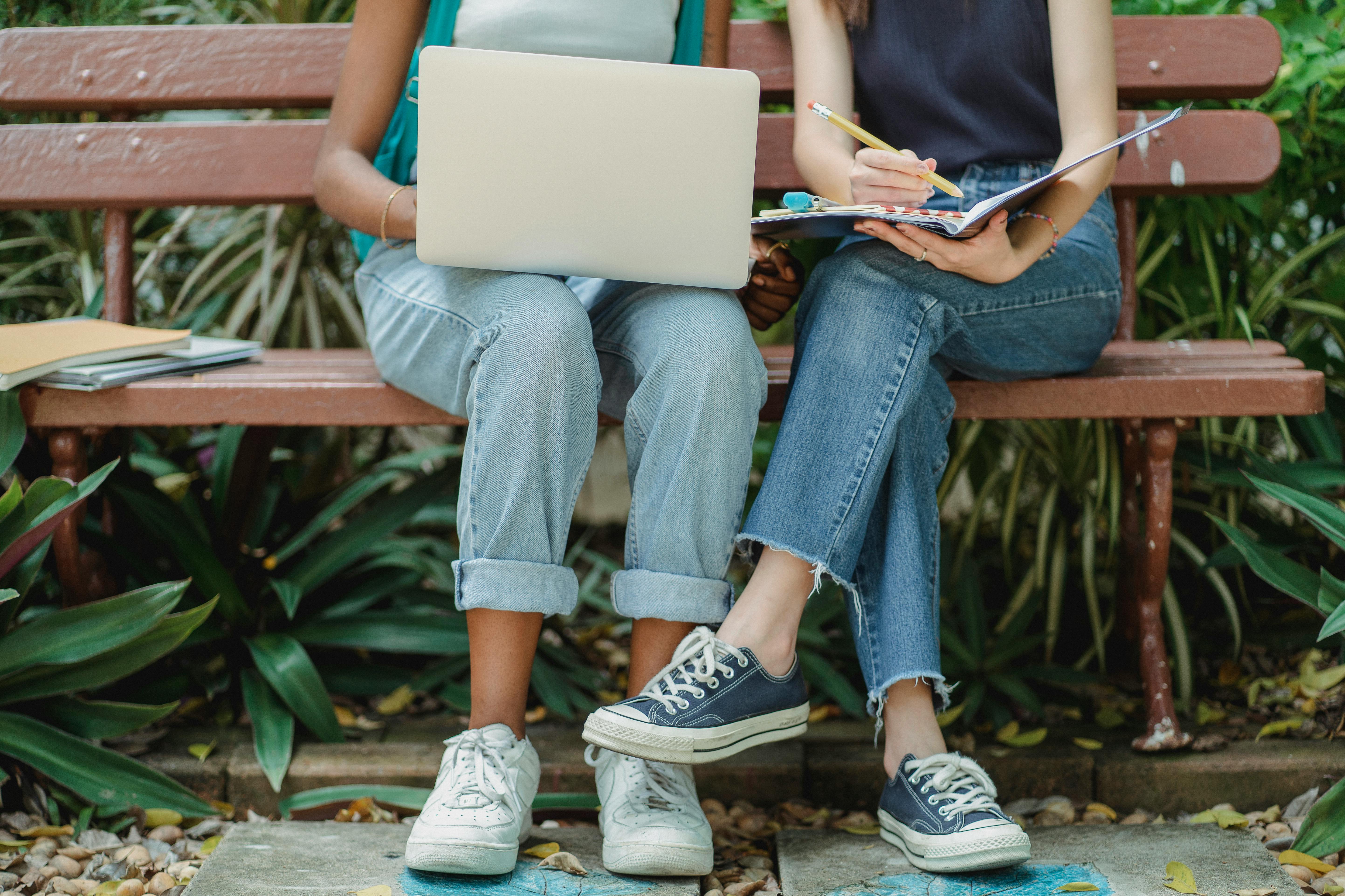 Two people wearing blue denim jeans sitting on a bench. The person sitting on the left-hand side is holding a computer and the other person is holding a notebook and a pencil.