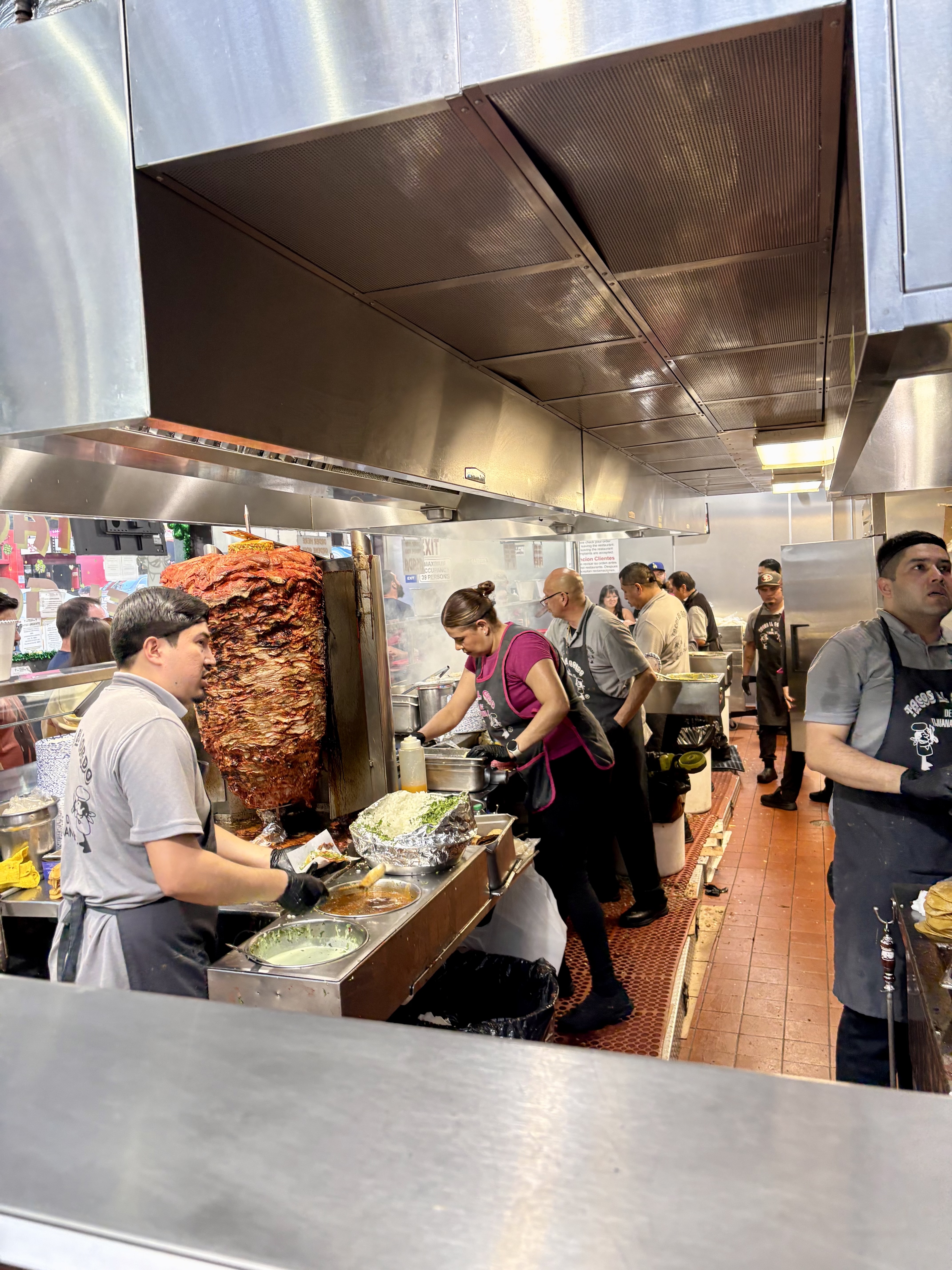 Inside the kitchen at Tacos El Gordo, showing staff working around a large trompo of rotating adobada pork.