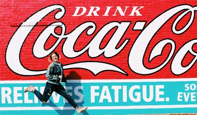 Amelia Kramer-Girl Jumping In Front Of Coca Cola Mural