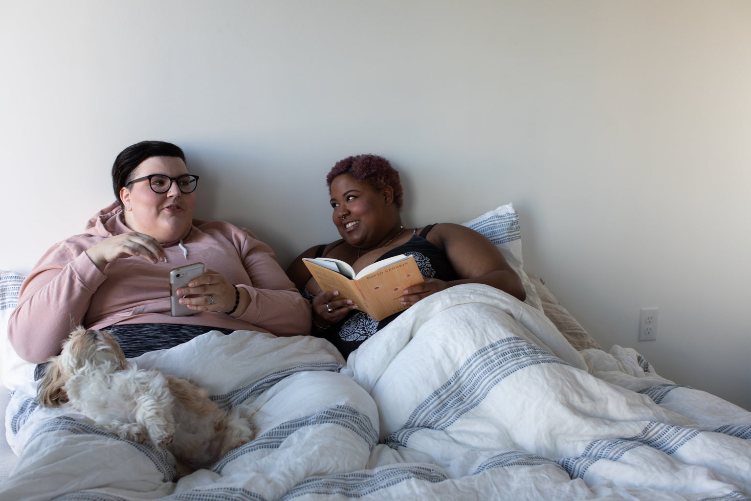 two plus size people in bed reading book and phone with dog