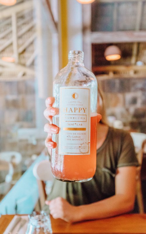 Girl holding up a kombucha bottle to camera covering her face