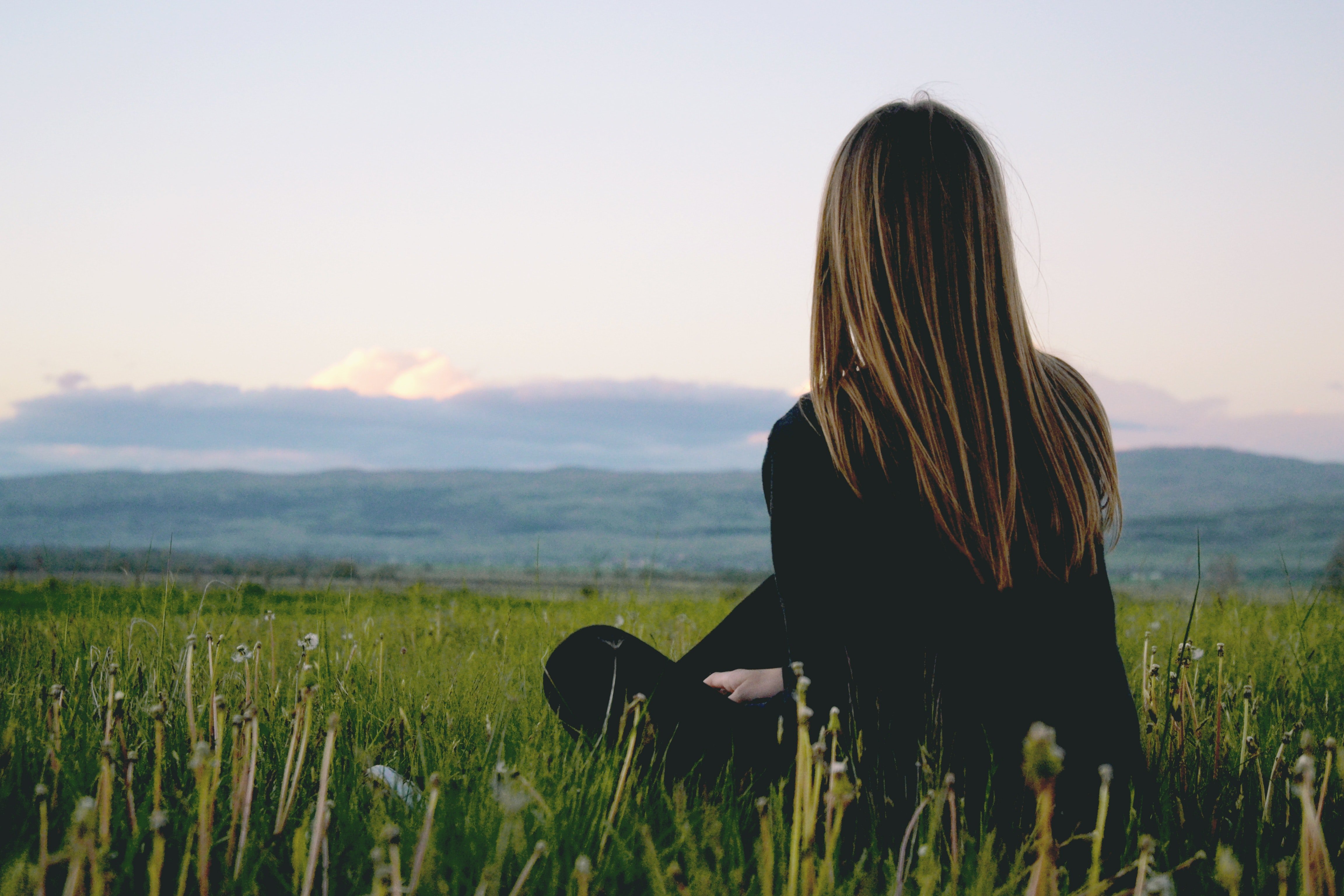 Woman wearing black long sleeved shirt sitting in green grass field near mountains under cloudy sky