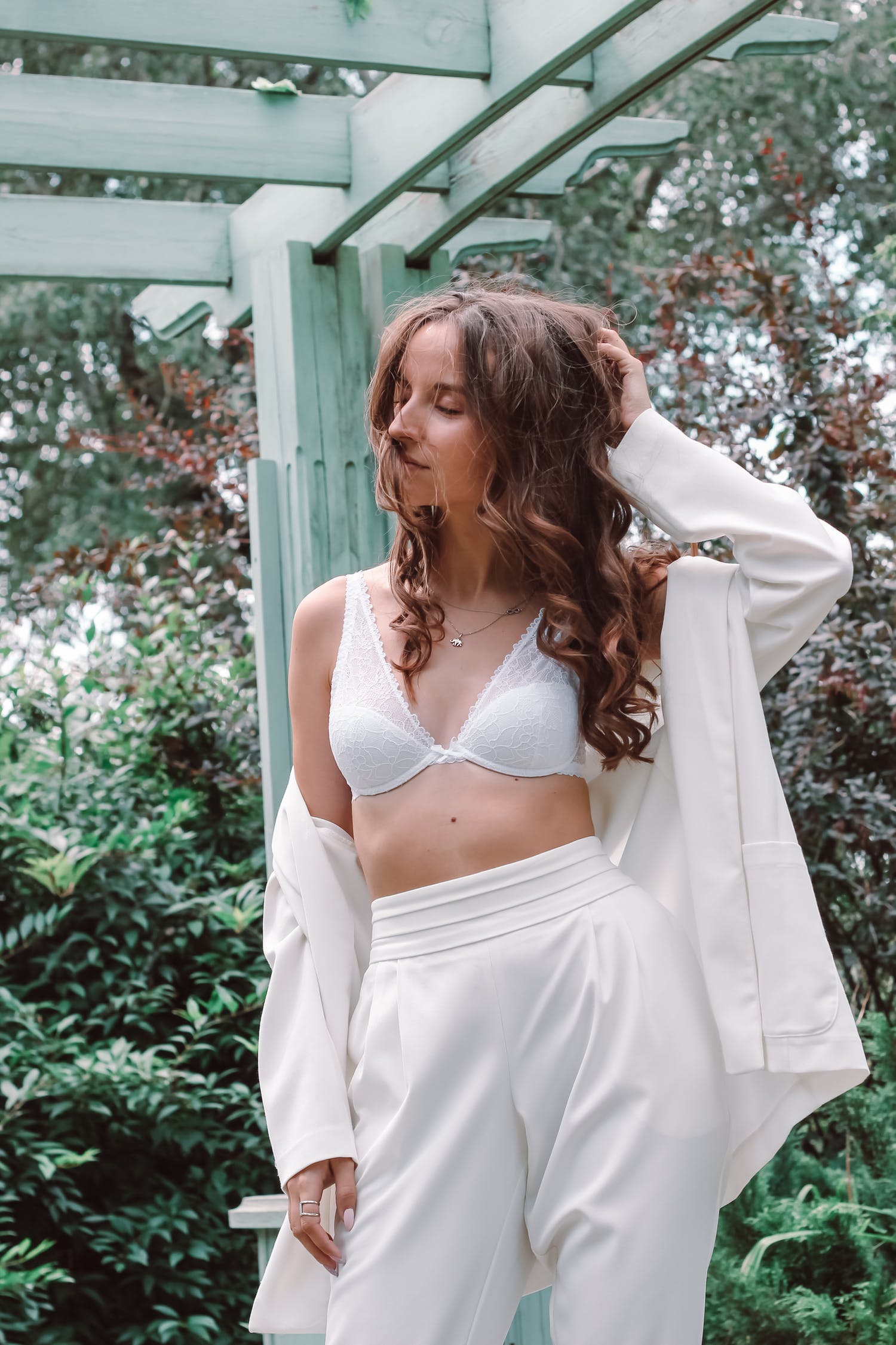 Woman in white suit and bralette standing near green plants.