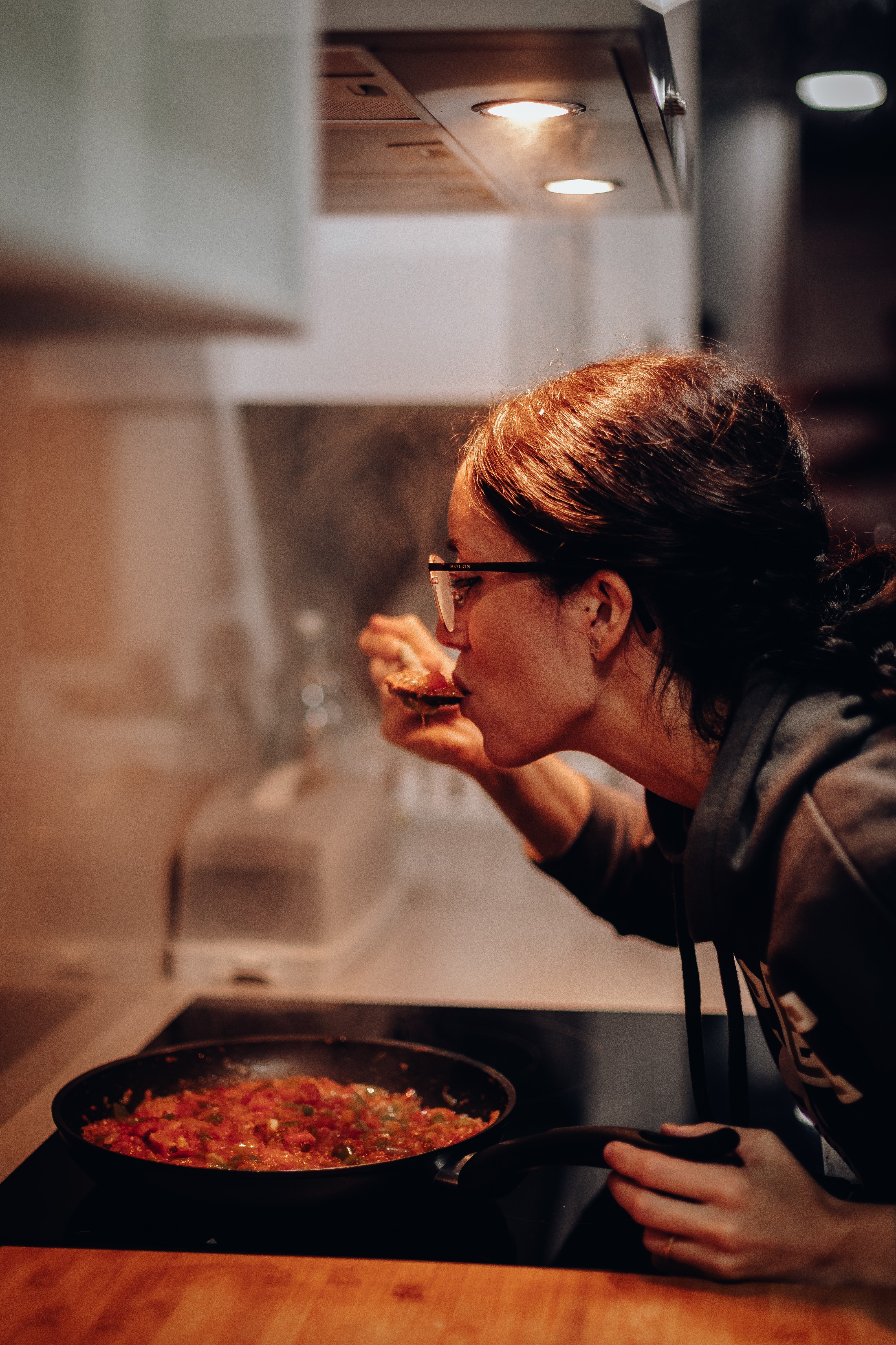 a woman leans over a pan of food, taste-testing it