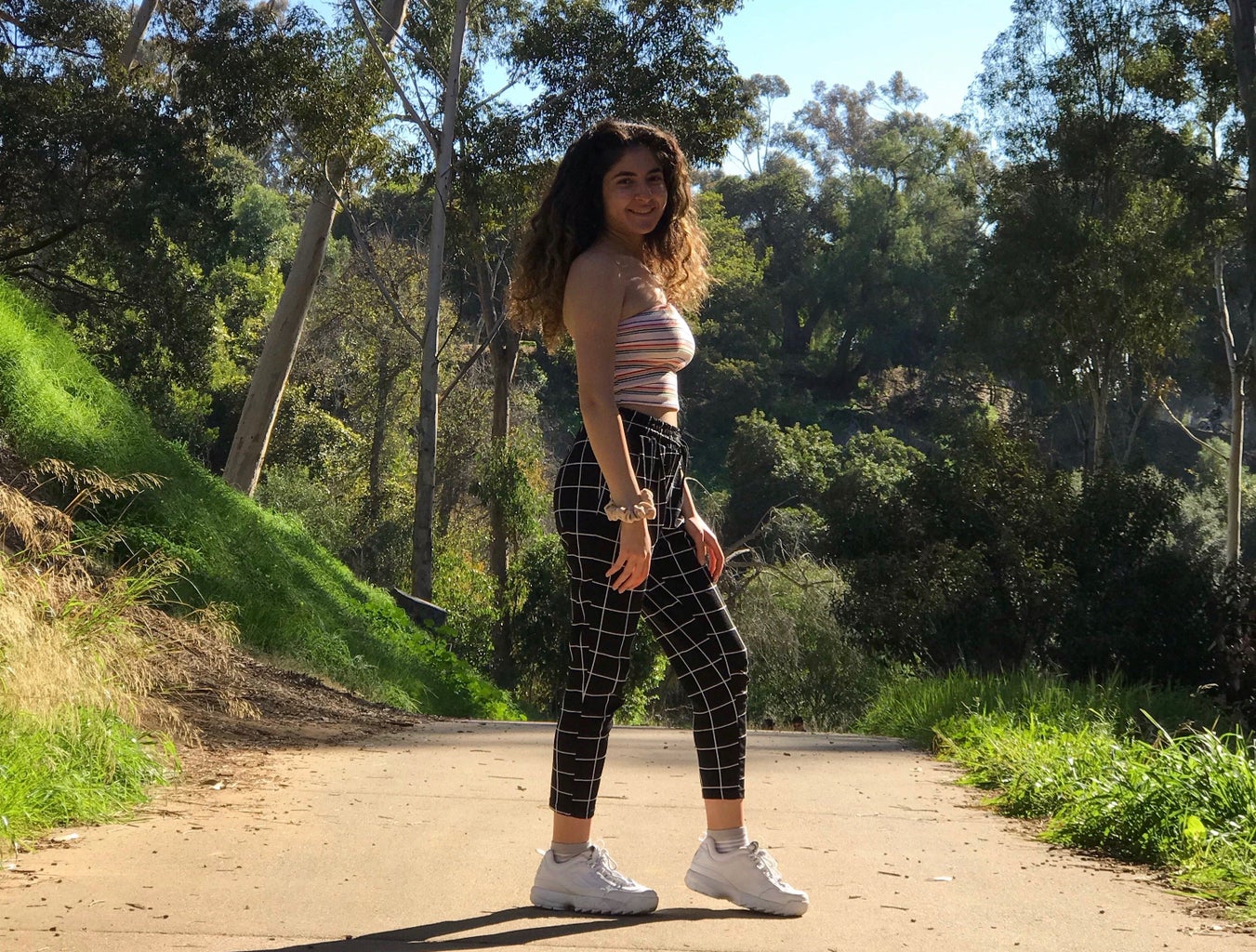 girl standing on a path with green trees around her; she is wearing grid pants and a striped tube top