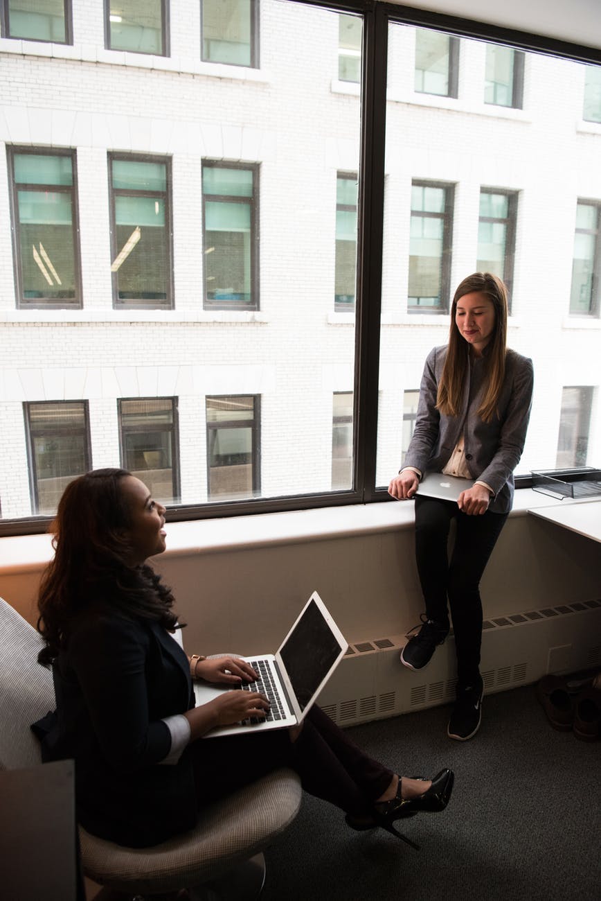 two women talking while holding laptops by the window at office
