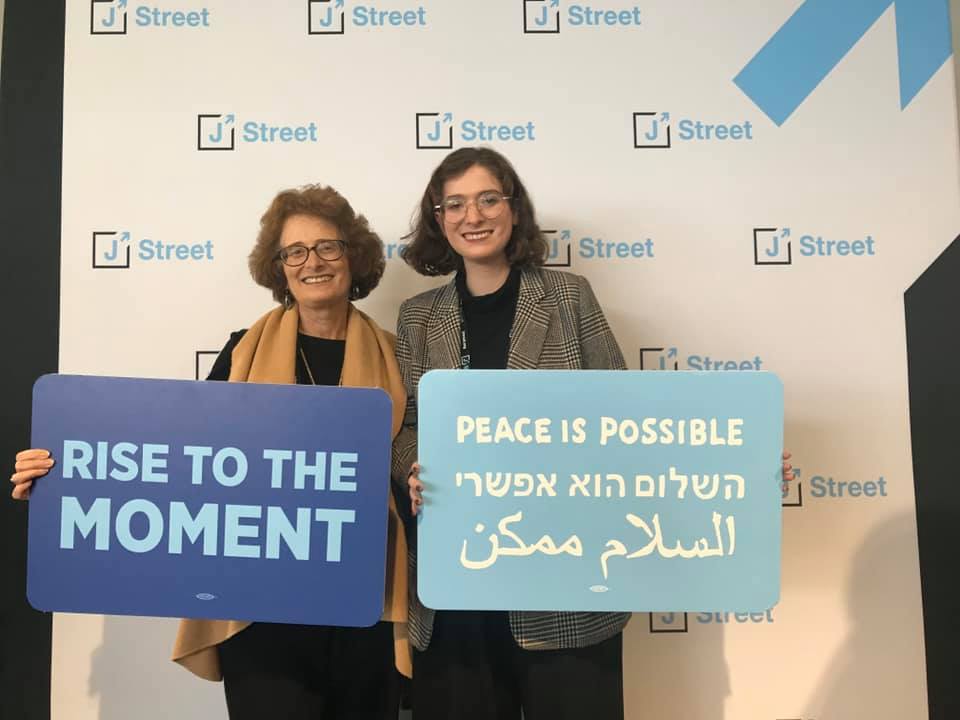Two women holding blue signs that say “rise to the moment” and “peace is possible”