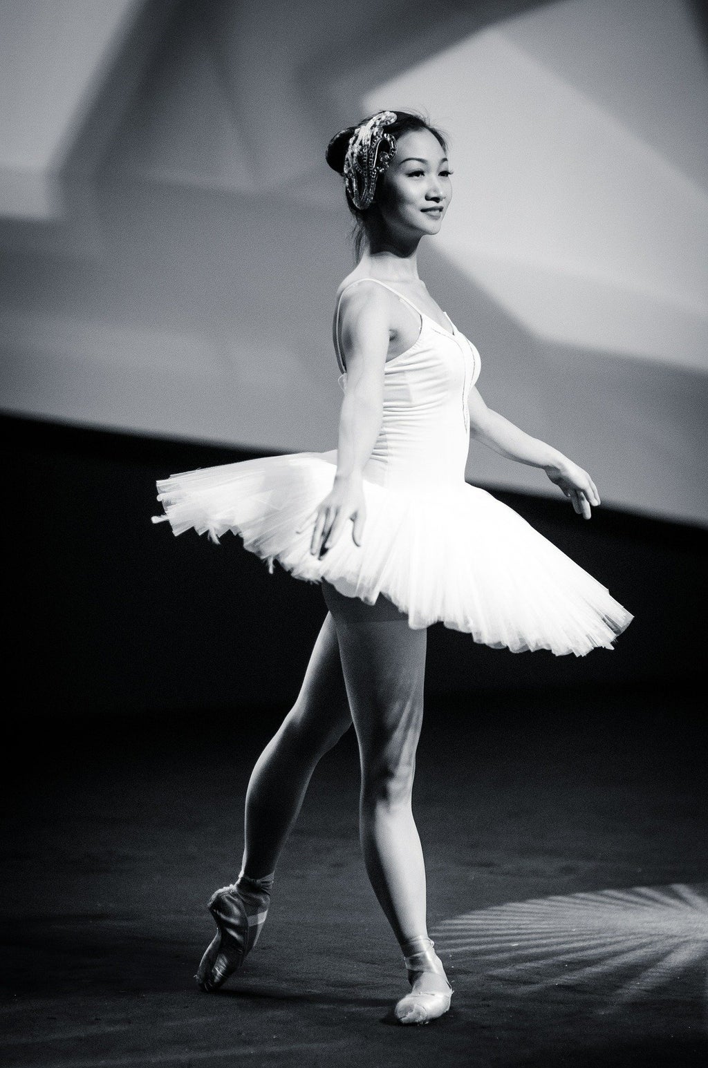 Black and white photo of woman wearing a white tutu, pointe shoes, and a decorative headpiece. Her arms are outstretched by her sides, and her left foot is extended behind her in the pointed tendu position.