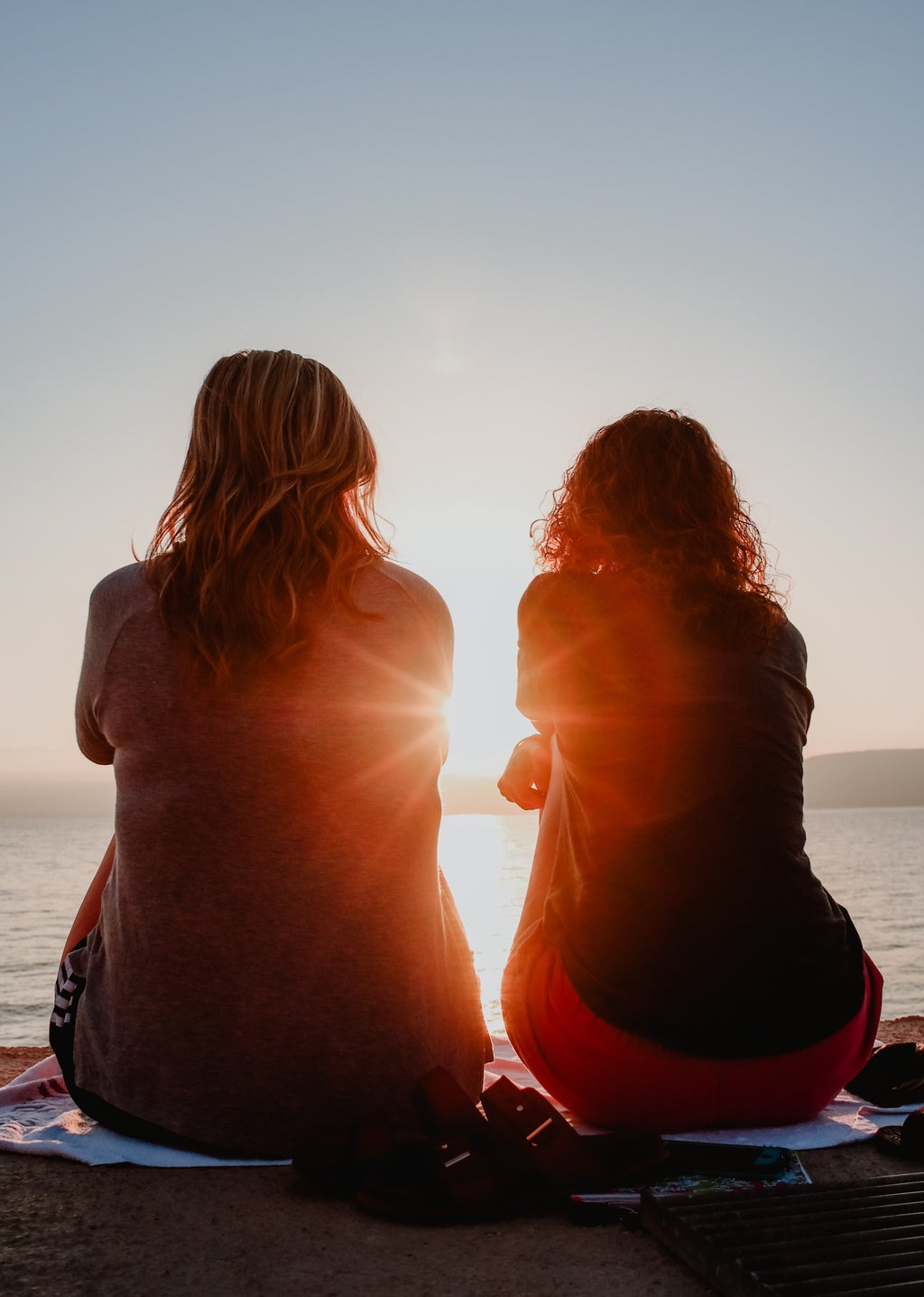 two women sit on the beach, facing the ocean. the sun shines in between them.
