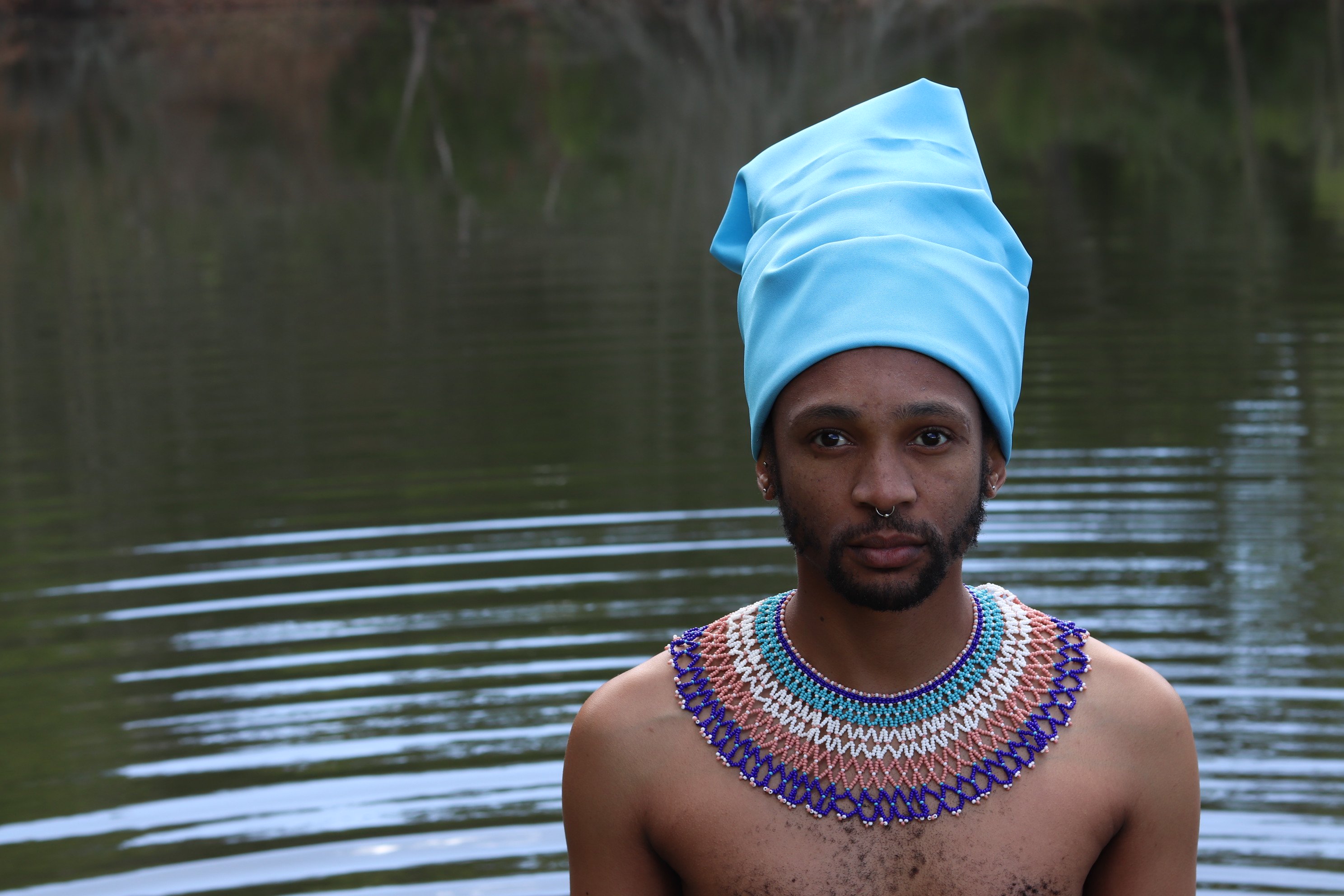 A Xhosa man, in traditional attire, stands infront of a body of water.