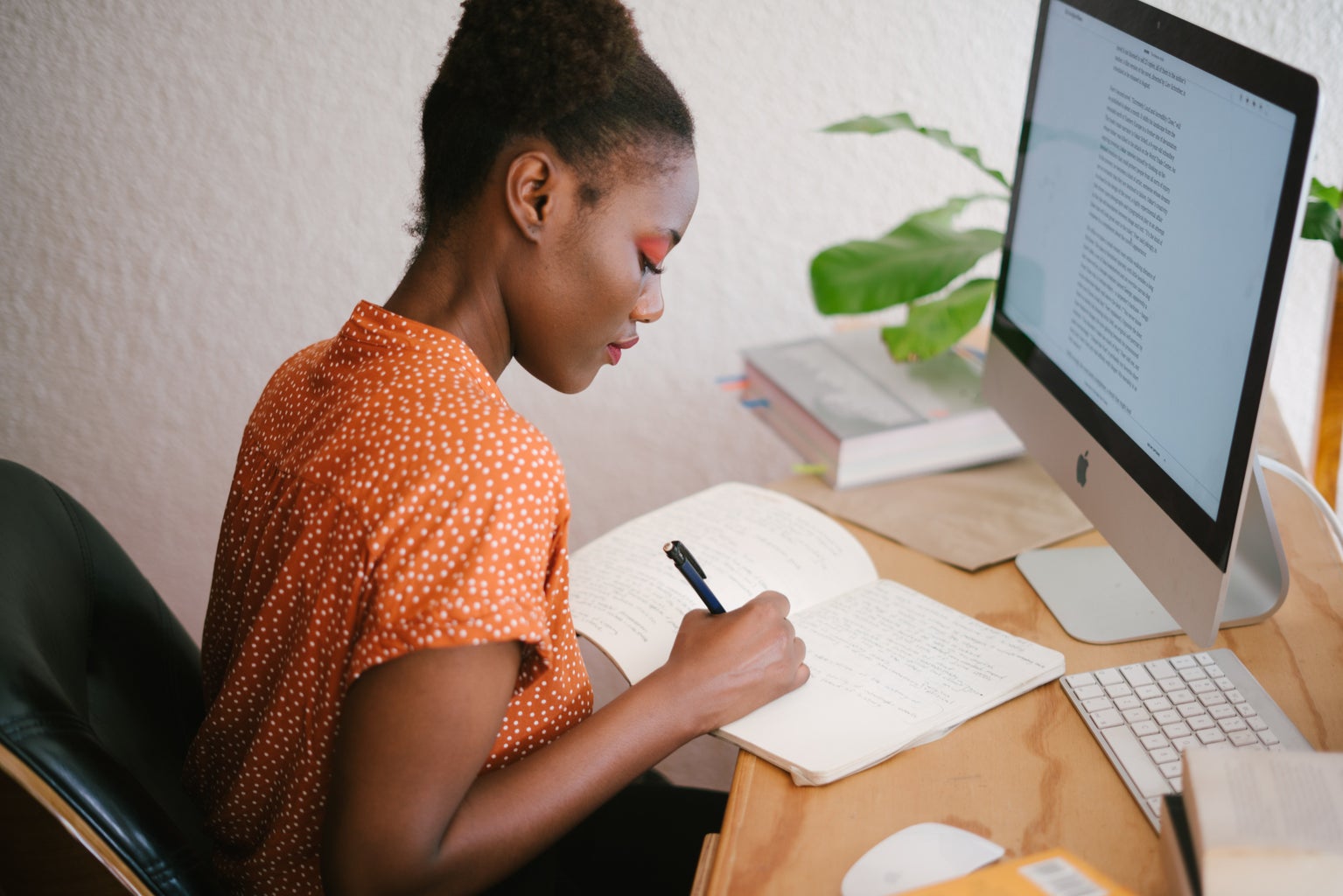 a woman sits at a wooden desk writing in a notebook. there is an imac in front of her.