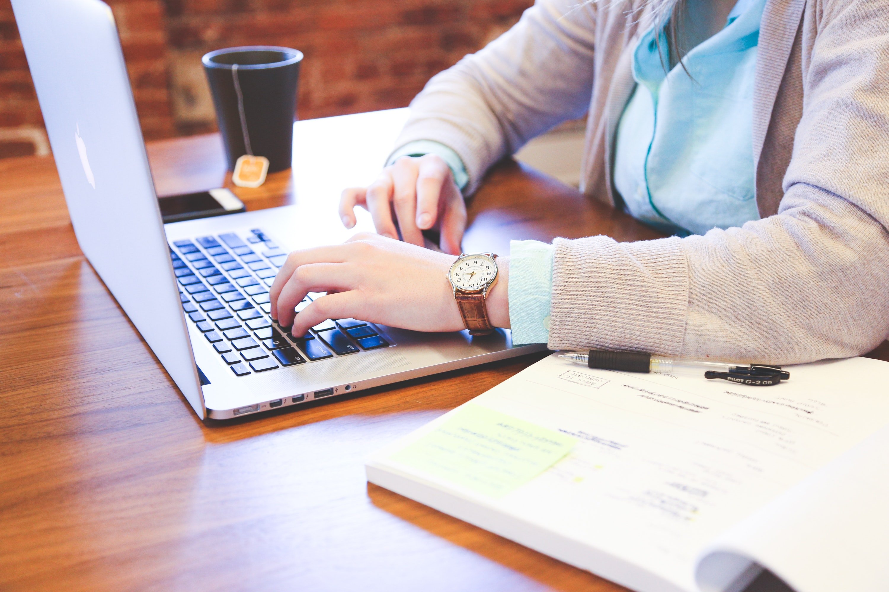 a person is seen sitting at a table, from the neck down. they are typing on a laptop and have a mug with tea and a highlighted book open next to them