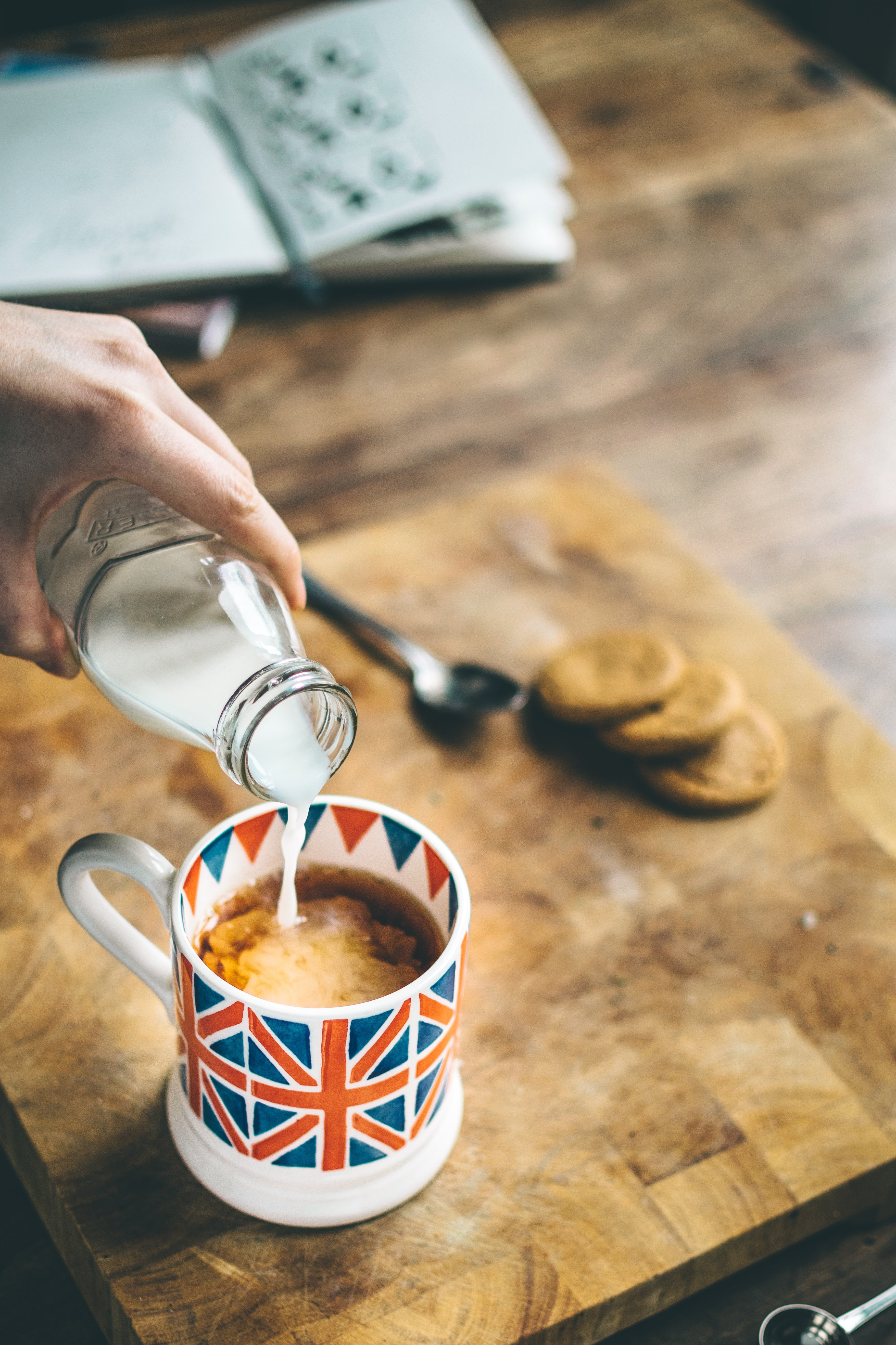 a hand is pouring milk out of a glass bottle into a cup of coffee, which is sitting on a wooden cutting board on a wooden table.