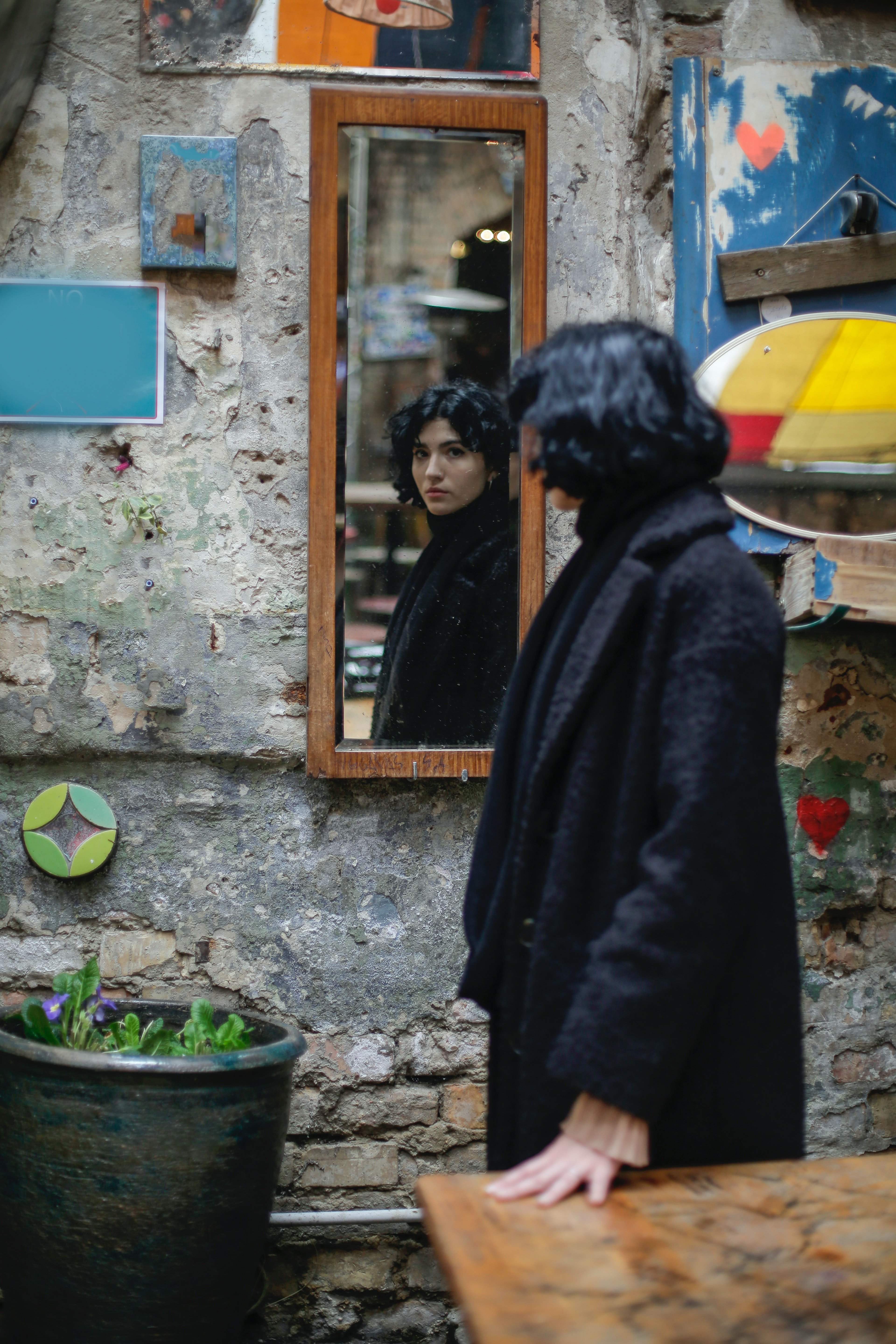 women in black coat near a brown wooden table looking in a mirror on the wall