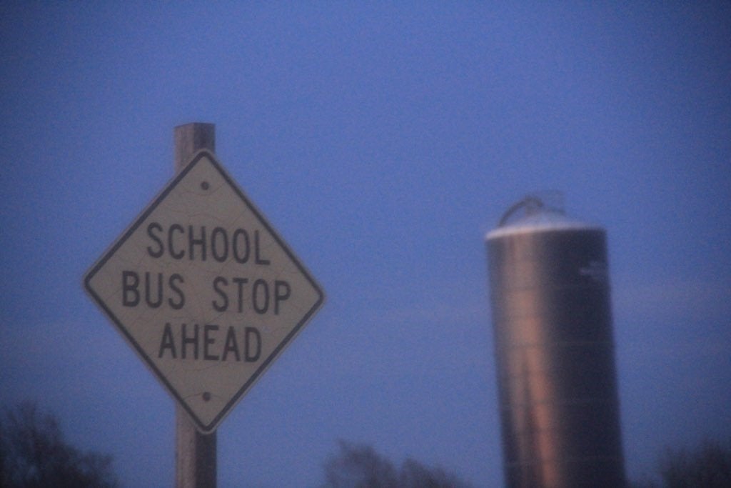 A \"school bus stopping zone\" sign with a farm building in the background.