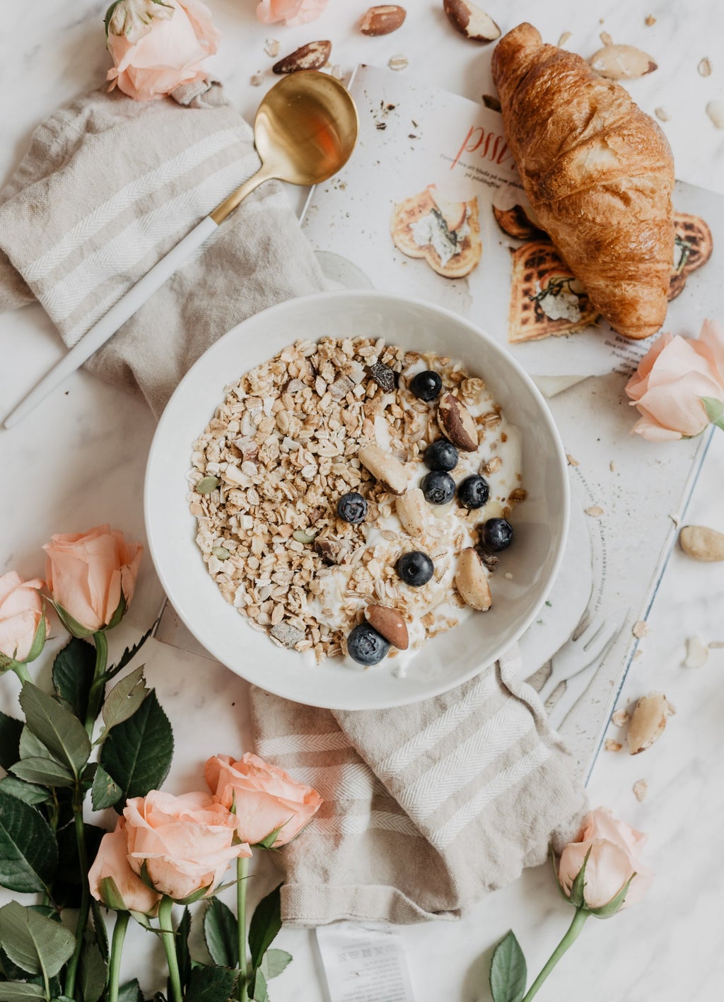 A lovely bowl of granola and berries surrounded by flowers and a croissant