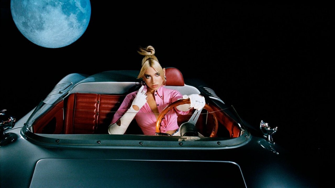Wide shot of Dua Lipa in a convertible with the moon in the upper left background