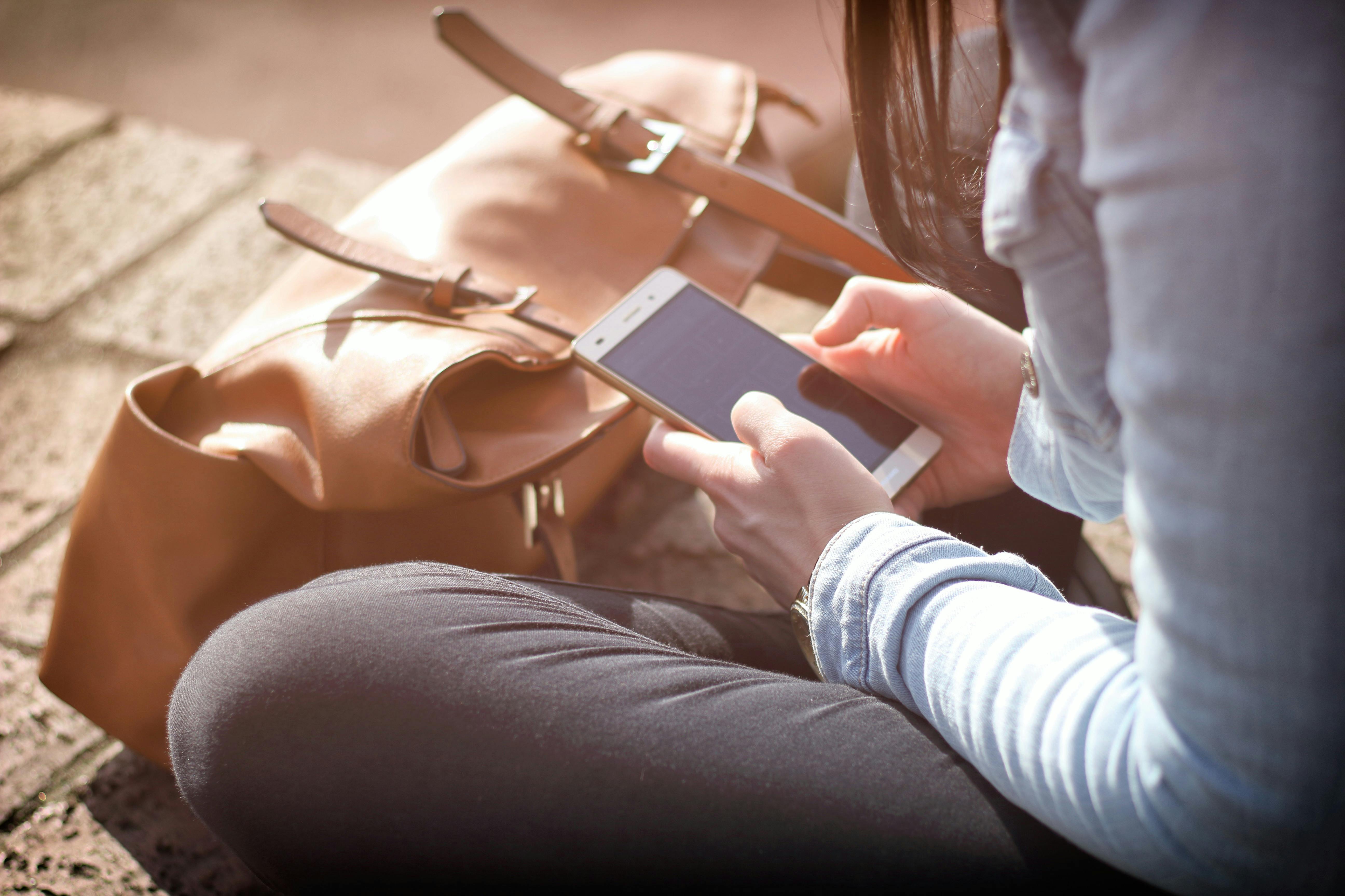women sitting on the sidewalk criss cross applesauce on her phone with her bag in front of her