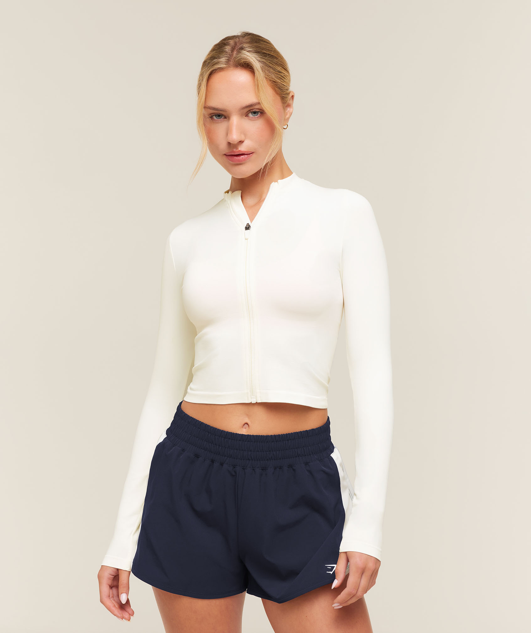 A blonde woman models a white fitted zip-up top and navy athletic shorts against a neutral studio backdrop, with a calm, confident expression.