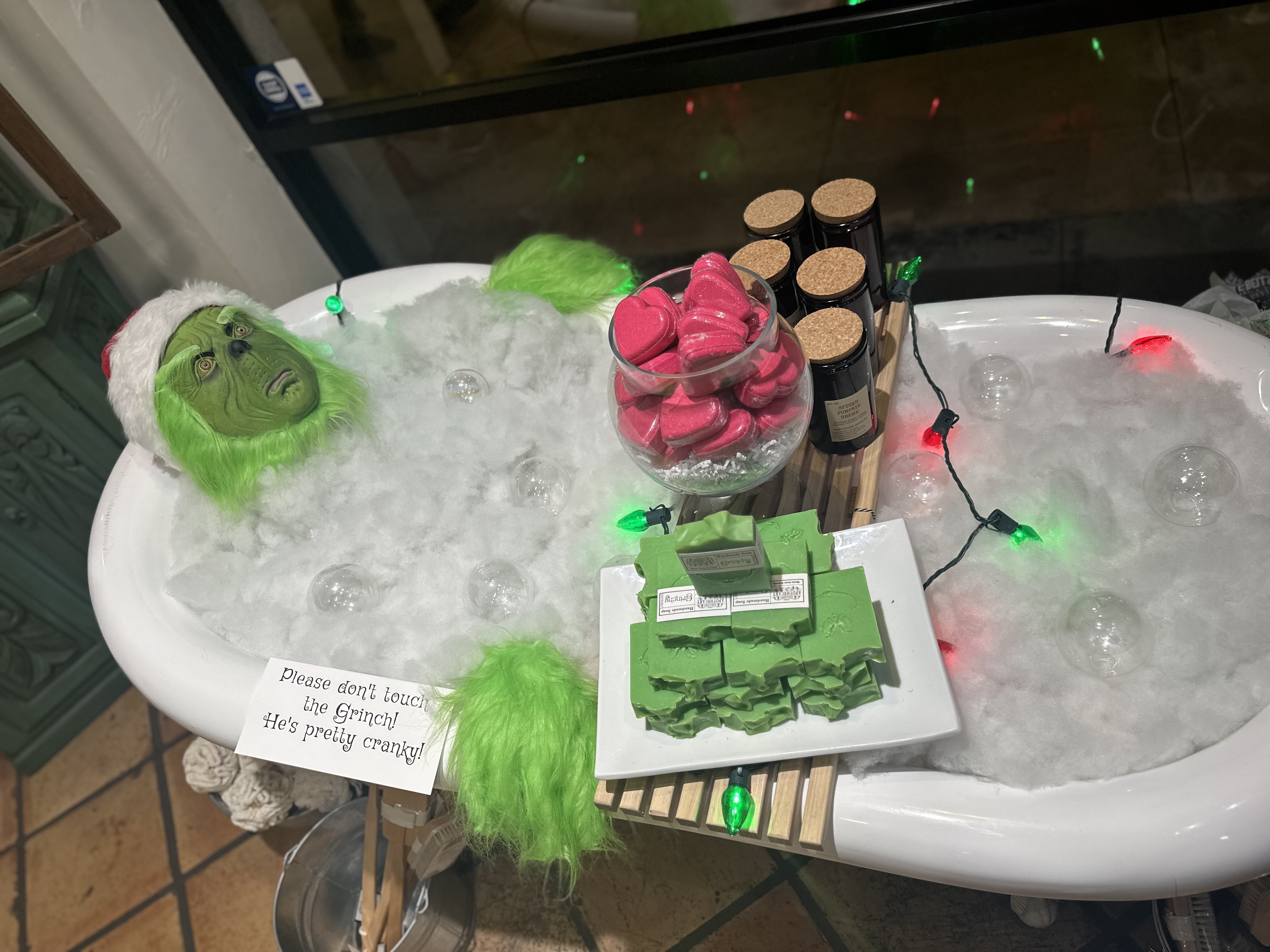 The Grinch with a Santa hat in a bath tub with fake soap and bubbles. Plate of soaps on top