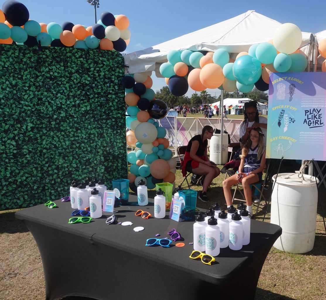 A photo of the Play Like A Girl tent, there is a photo wall that is green and balloons that are teal and orange with tables in front  that have water bottles on display