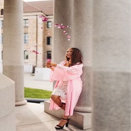 Young woman in the pink graduation gown popping a bottle of champagne with confetti against the marble columns background.