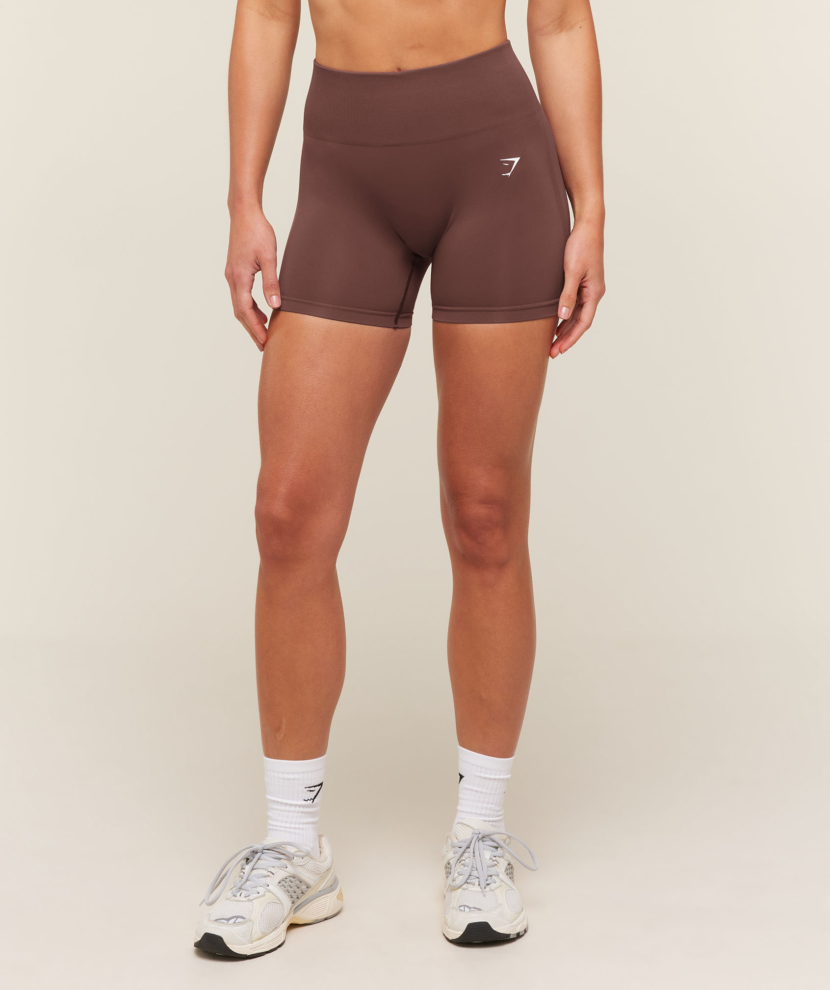 A model stands against a neutral studio backdrop wearing dark brown fitted athletic shorts, white crew socks, and running shoes, showcasing the workout apparel.