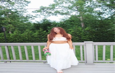 Young female with long reddish colored hair standing in a white two piece set standing on a deck with green trees in the background