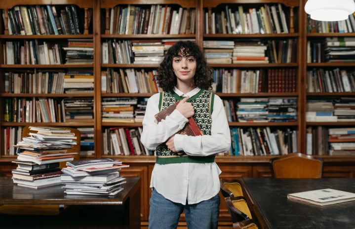 Woman in sweater vest stands in messy library.