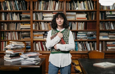 Woman in sweater vest stands in messy library.