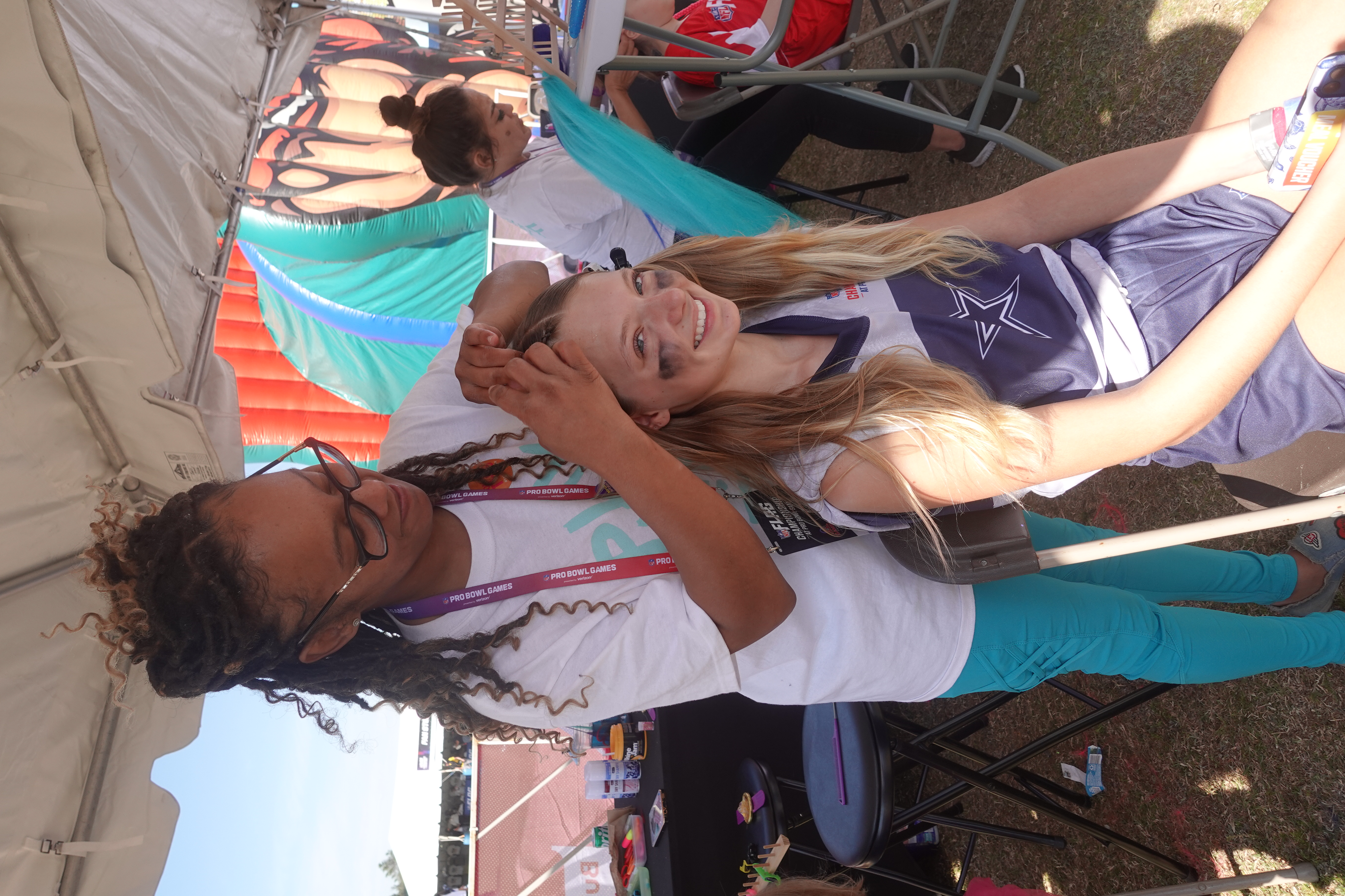 An image of a young girl with a Cowboys logo on her jersey smiling while getting her hair braided under a tent