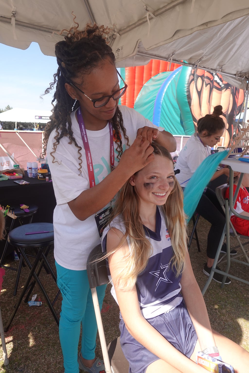 An image of a young girl with a Cowboys logo on her jersey smiling while getting her hair braided under a tent