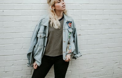 Woman in blue denim jacket and black pants standing beside gray wall