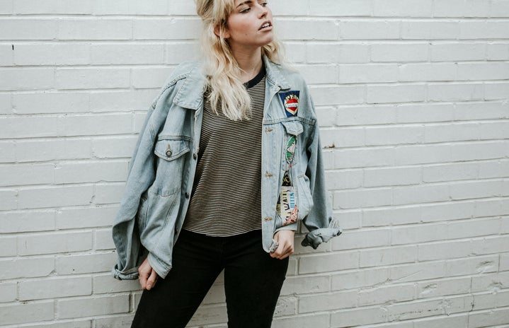 Woman in blue denim jacket and black pants standing beside gray wall