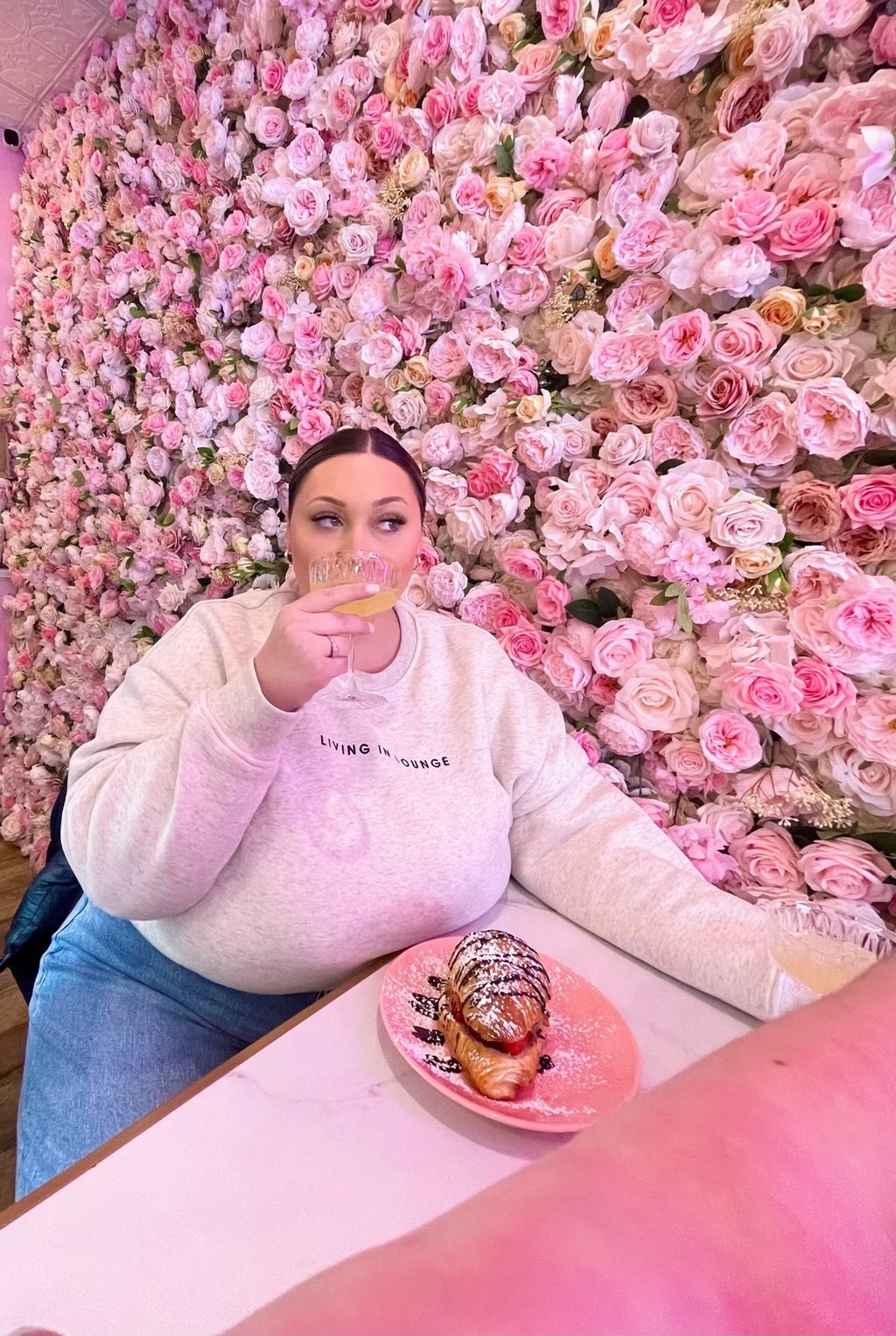 Woman drinking a glass of juice in front of a flower wall.