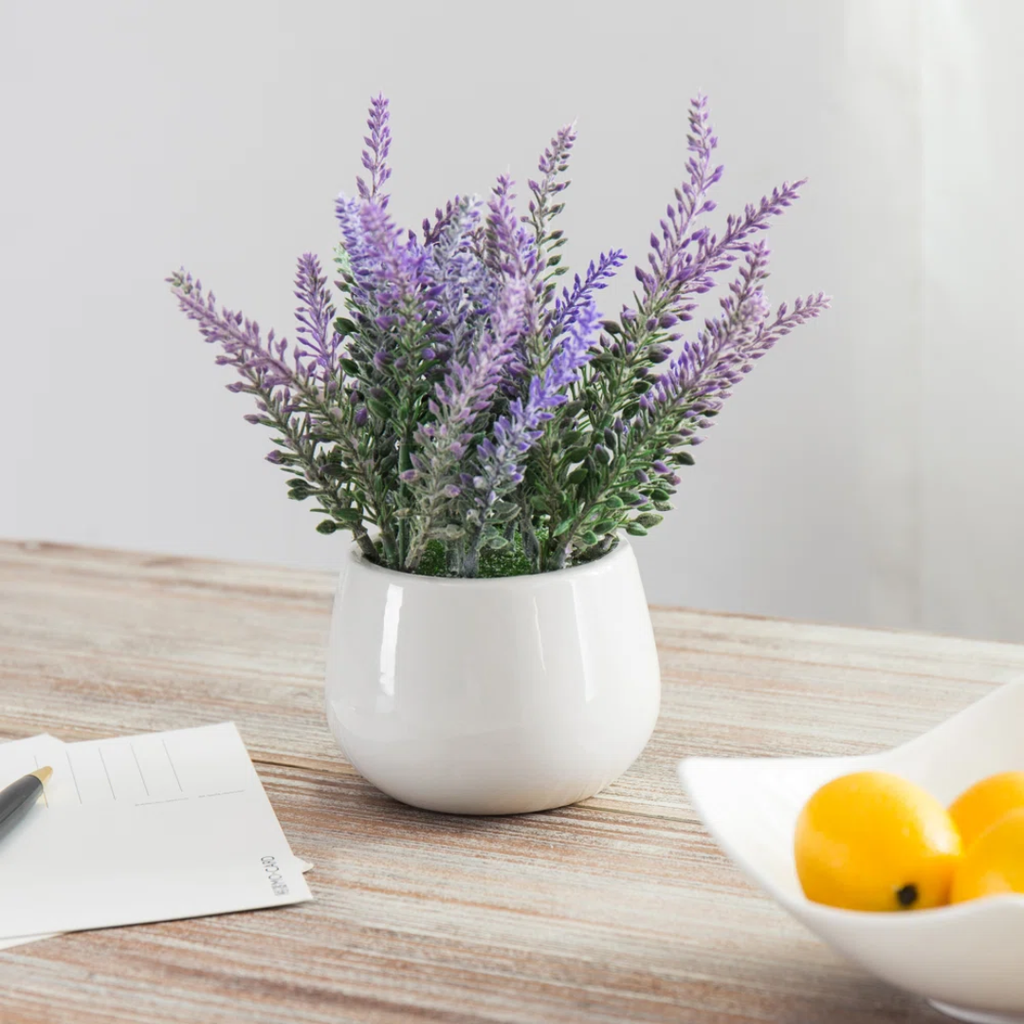 faux lavender plant in white pot on table next to a piece of paper and a bowl of oranges