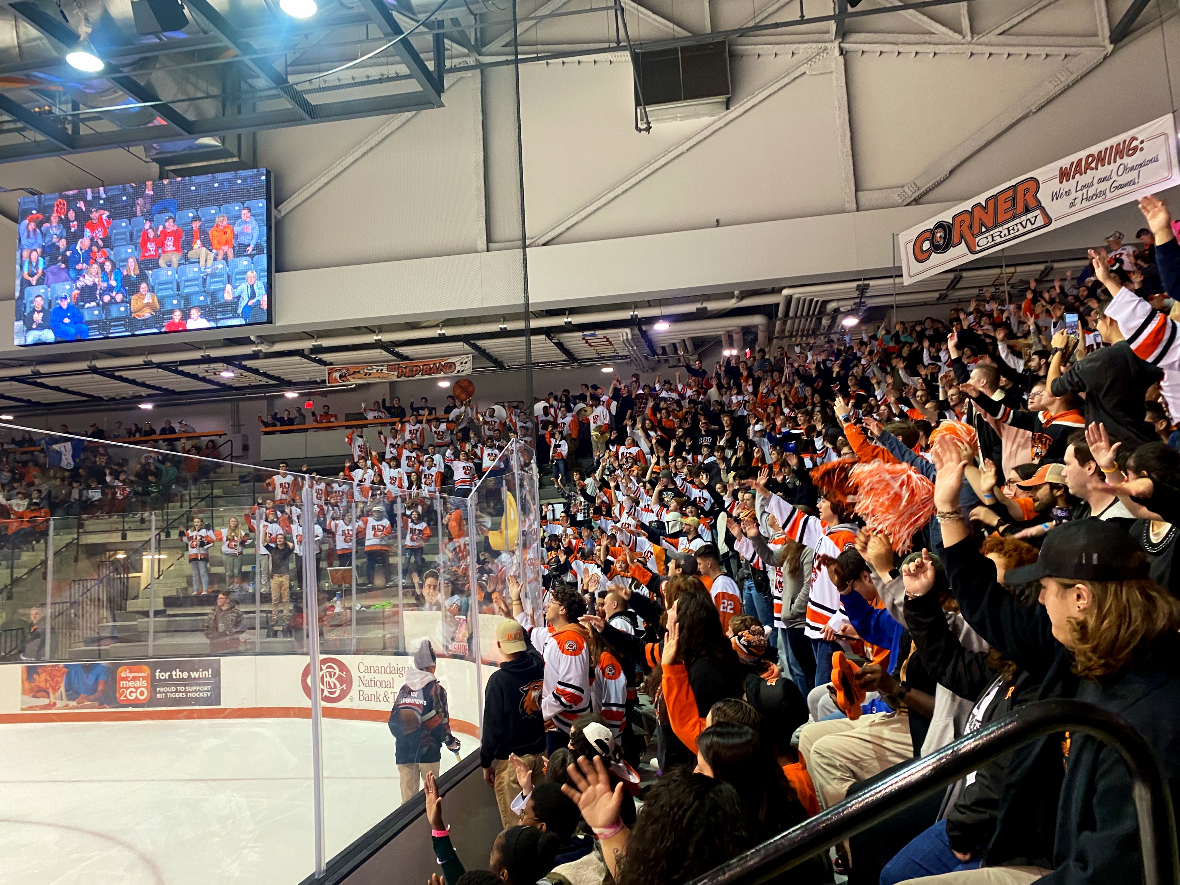 RIT Men;s Hockey team: audience cheering for team in seating row, wearing orange, white, and black.