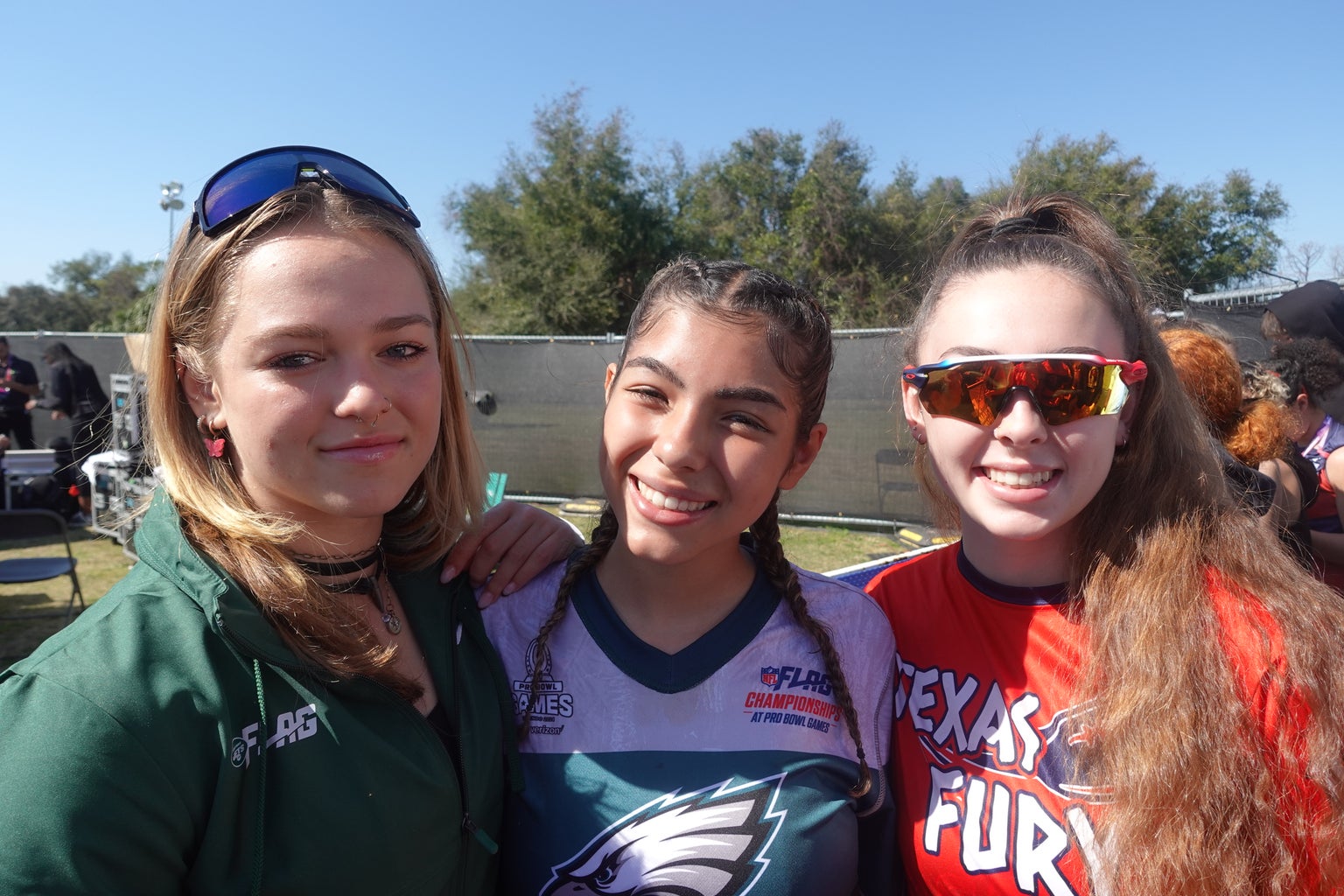 A photo of 3 17u girls flag football players. From left to right a blonde with a green sweater, a brunette with braids and an eagles jersey and a brunette with sunglasses and a red \