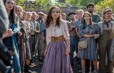 Lucy Gray Baird in a purple skirt and white top surrounded by civilians in the film, \"The Hunger Games: The Ballad of Songbirds and Snakes\"