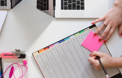 Person writing on sticky note with planner and laptop in background